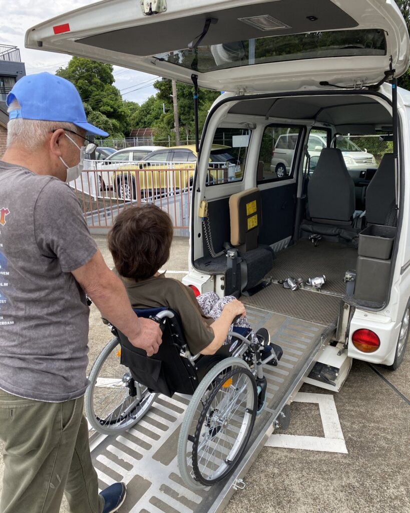 Driver assisting a passenger in a wheelchair up the rear ramp of our compact van, with floor securement rails visible